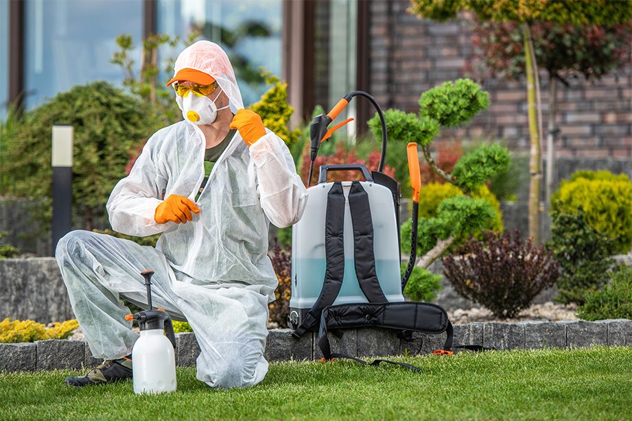 TCB Pest Control technician in PPE preparing a backpack sprayer beside a garden bed